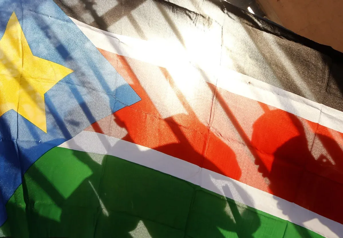 علم جنوب السودان يتعرض لأشعة الشمس، مع ظلال لأشخاص يحملون أعلامًا، مما يعكس الوضع السياسي والفساد في البلاد.