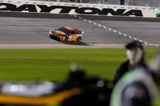 سيارة سباق تتجه نحو خط النهاية في دايتونا بيتش، مع وجود حلبة السباق في الخلفية، تعكس أجواء المنافسة في رياضة ناسكار.