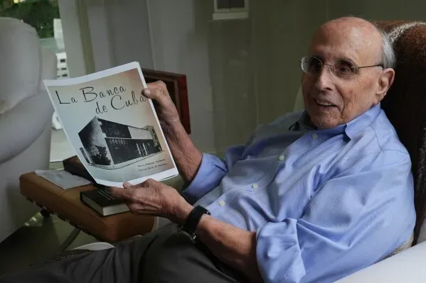 رجل مسن يجلس في غرفة معيشته، يحمل كتيبًا بعنوان "La Banca de Cuba"، مع خلفية تضم كتبًا وصورًا تعكس تاريخ كوبا.