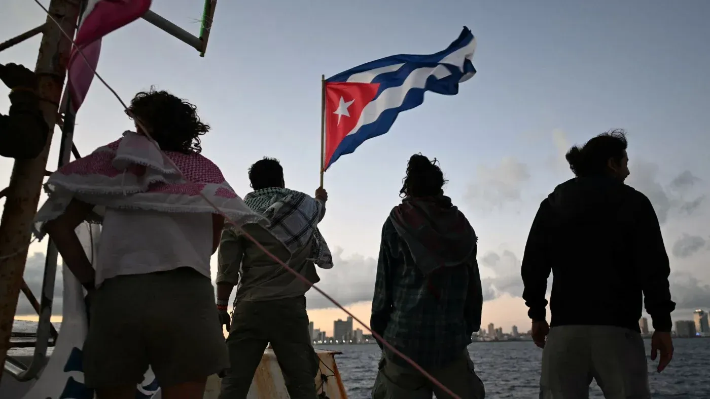 مجموعة من الأشخاص يقفون على ضفاف البحر في هافانا، يحمل أحدهم العلم الكوبي، مع خلفية المدينة في الغسق، تعبيرًا عن التضامن.