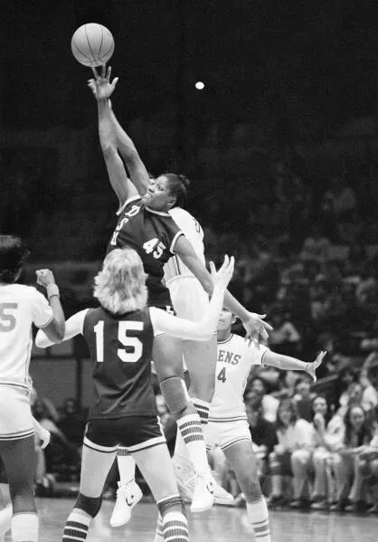 لاعبة كرة سلة ترتفع لتسديد الكرة خلال مباراة في السبعينيات، مع مشجعات في الخلفية، تعكس بداية تاريخ كرة السلة النسائية في ولاية دلتا.