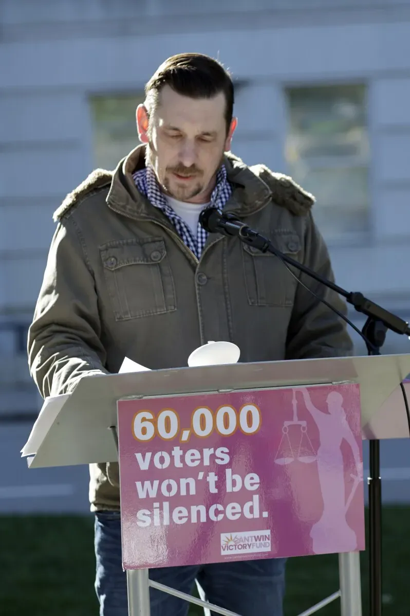 رجل يتحدث خلف منصة تحمل لافتة مكتوب عليها "60,000 ناخب لن يتم إسكاتهم"، يشير إلى قضايا حقوق الناخبين في نورث كارولينا.