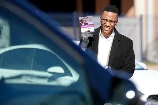 كريستيان منفي، المرشح لمجلس النواب الأمريكي، يحمل منشورًا انتخابيًا مبتسمًا أثناء حملته في هيوستن، مع سيارات خلفه.