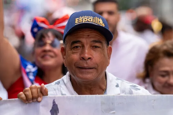 رجل يحمل لافتة في مسيرة، يرتدي قبعة تحمل اسم "Adriano Espaillat"، ويظهر خلفه حشد من المشاركين في الاحتفال.