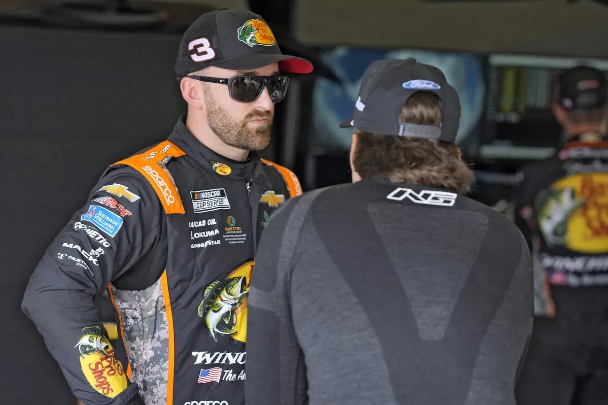 سائق NASCAR يرتدي زي السباق ويتحدث مع زميله في الحلبة، مع خلفية تظهر سيارات سباق أخرى، خلال التجارب الحرة لسباق دايتونا 500.