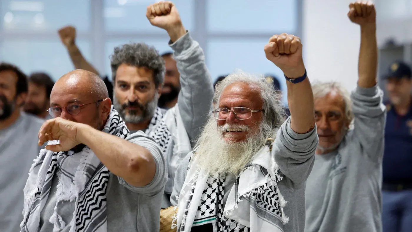 ناشطون يرفعون قبضاتهم في مطار إسطنبول بعد الإفراج عنهم من الاحتجاز الإسرائيلي، يرتدون بدلات السجن الرمادية والكوفية.