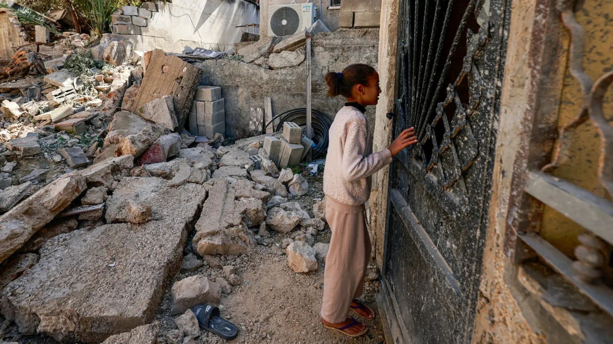طفلة فلسطينية تقف أمام باب منزل مدمر في جنين، محاطة بأنقاض منازل مهدمة، تعكس آثار الهجوم العسكري المستمر.
