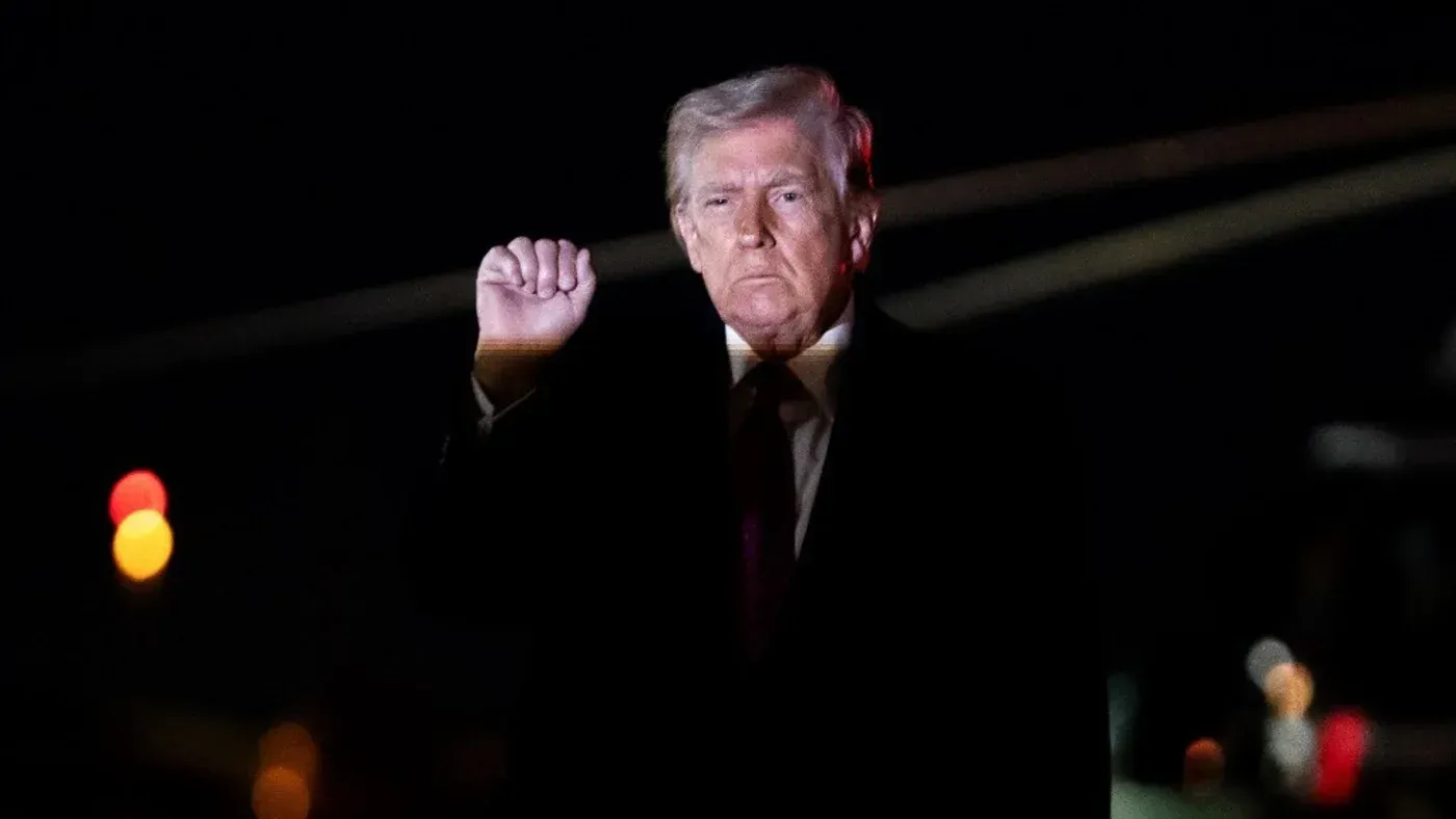 ترامب يرفع يده بإشارة قوية، مع خلفية مضاءة خافتة، معبرًا عن مواقفه حول الاستيلاء على النفط الإيراني.