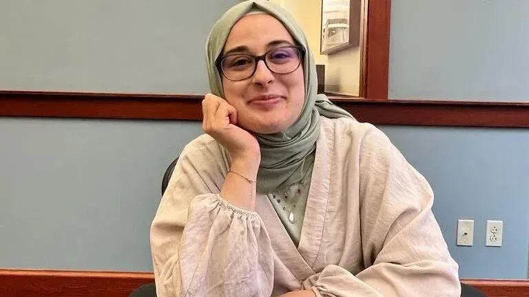 طالبة ترتدي حجابًا وتبتسم في غرفة مكتب، تعبيرها يوحي بالثقة، في سياق اعتقالها بسبب آرائها المؤيدة لفلسطين.