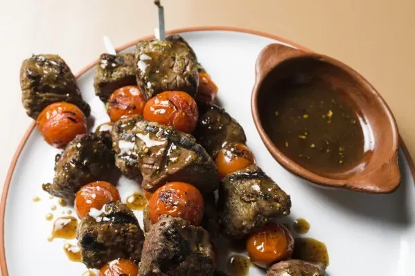 أسياخ لحم البقر المشوي مع الطماطم، مغطاة بصلصة ماديرا، تقدم على طبق مع وعاء جانبي يحتوي على الصلصة، تعكس نكهات الوصفة البرتغالية التقليدية.