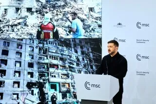 رجل يتحدث أمام منصة في مؤتمر أمني، مع صورة خلفه تظهر دمار مبنى سكني نتيجة الهجمات في أوكرانيا.