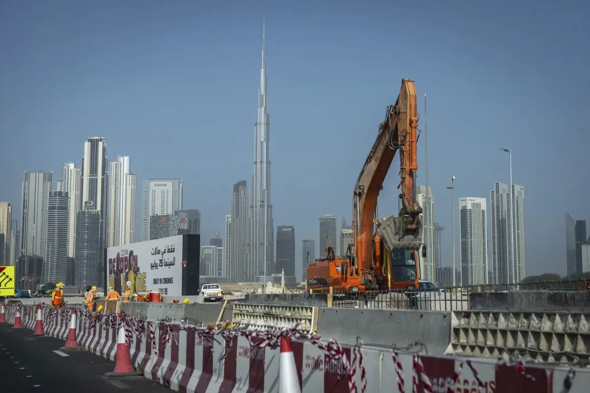 تظهر الصورة عمالاً في موقع بناء بالقرب من ناطحات السحاب في دبي، مع برج خليفة كخلفية، مما يعكس النمو السريع والتطور العمراني في المدينة.