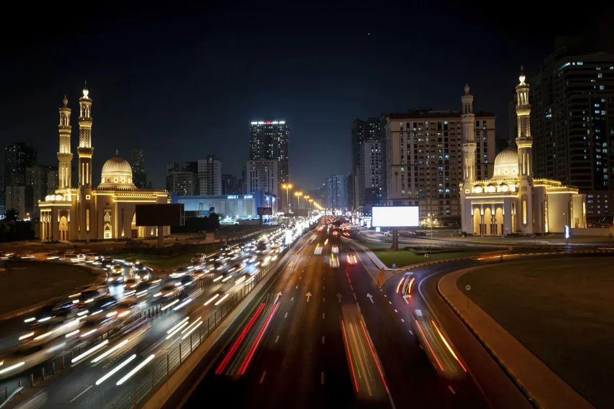 تظهر الصورة مشهداً ليلياً لطرق دبي مزدحمة بالمركبات، مع معالم معمارية مضيئة على الجانبين، تعكس النمو الحضري في المدينة.