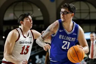 لاعب كرة سلة يرتدي نظارات، يتجاوز مدافعًا أثناء مباراة بين فريق بيليكنز وديفيدسون، مع التركيز على الكرة.