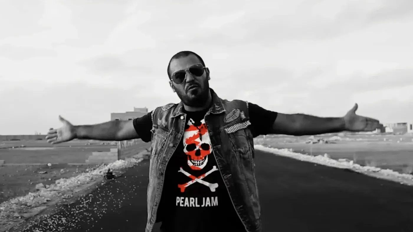 مغني راب ليبي يرتدي سترة جلدية وقميص يحمل شعار "Pearl Jam"، يمد ذراعيه في مشهد يعكس حرية التعبير وسط القيود المفروضة.
