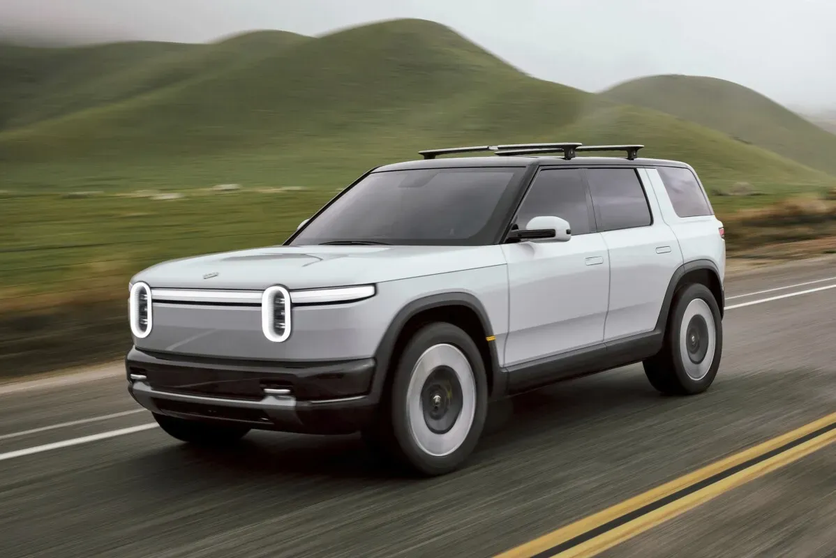 سيارة Rivian R2 الرياضية متعددة الاستخدامات تسير على طريق محاط بالتضاريس الخضراء، مع تصميم عصري ومظهر متين.