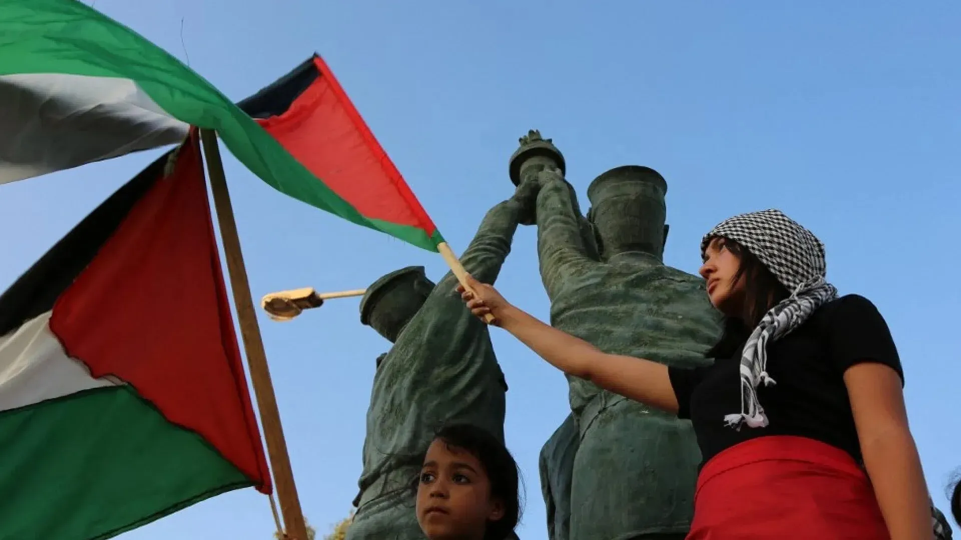 امرأة وطفلة تحملان علم فلسطين أمام تمثال كبير، تعبيرًا عن التضامن مع غزة في المسيرة العالمية.