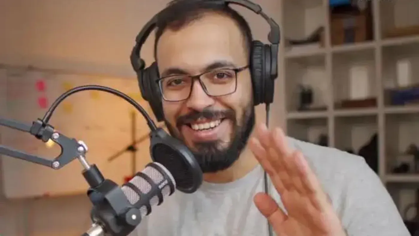أحمد أبو زيد، مستخدم يوتيوب شهير، يتحدث في البودكاست مع ميكروفون، ويظهر خلفه أدوات تصوير، مع التركيز على محتواه التعليمي.