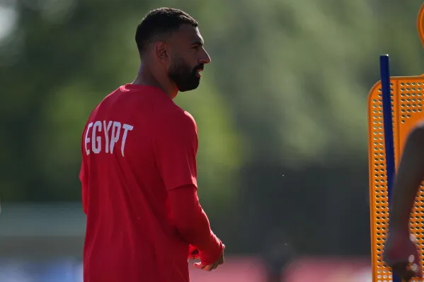 لاعب مصري يرتدي قميصًا أحمر مكتوب عليه "مصر"، يستعد للتدريب قبل مباراة كأس العالم ضد نيوزيلندا، مع التركيز على تحقيق إنجاز تاريخي.