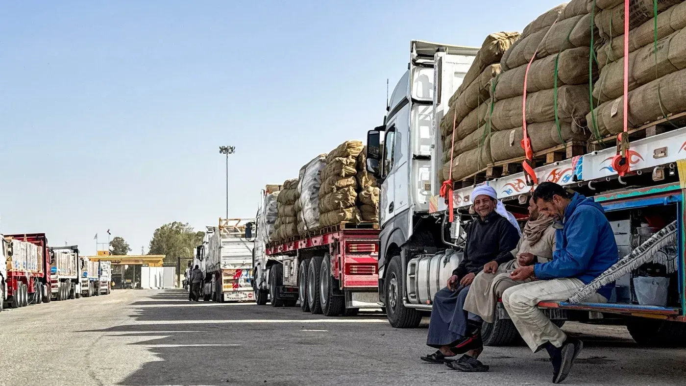 شاحنات محملة بالمساعدات الإنسانية متوقفة عند معبر رفح، مع وجود رجال يجلسون بالقرب منها، استعدادًا لإدخال المساعدات إلى غزة.