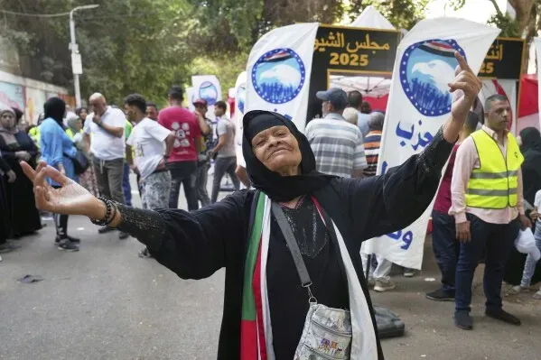 امرأة مسنّة ترتدي زيًا تقليديًا وتحتفل أمام مركز اقتراع في القاهرة، وسط حشود من الناخبين ولافتات انتخابية لمجلس الشيوخ.