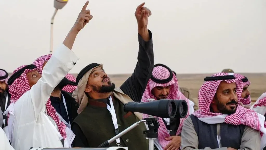 مجموعة من الرجال يرتدون الملابس التقليدية السعودية، يستخدمون تلسكوبات للتحري عن رؤية الهلال في السماء، مع تعبيرات من التركيز والاهتمام.