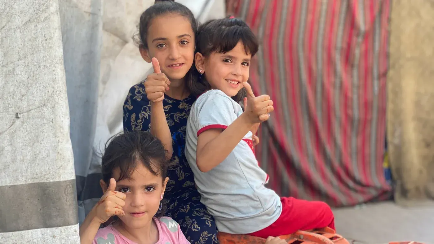 ثلاث فتيات صغيرات يجلسن معًا في مكان مفتوح، يبتسمن ويرفعن أصابعهن بإشارة الإعجاب، في أجواء عيد الفطر بغزة.