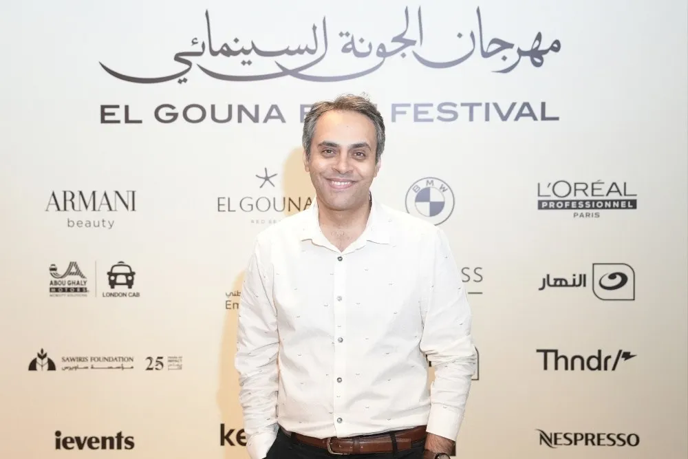 أندرو محسن مدير فني جديد لمهرجان الجونة السينمائي مبتسم أمام لافتة المهرجان التي تبرز دعم الأفلام العربية والأفريقية.