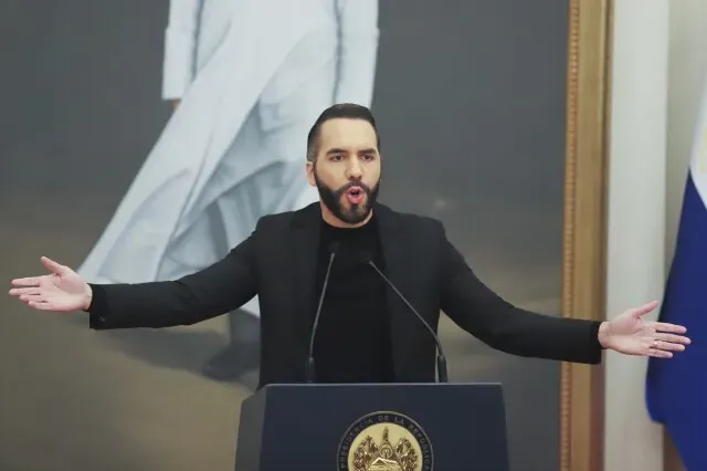 الرئيس ناييب بوكيلي يتحدث بحماس خلال مؤتمر صحفي، مع وجود علم السلفادور في الخلفية. يناقش تمديد فترة الحبس الاحتياطي للمتهمين بالارتباط بالعصابات.