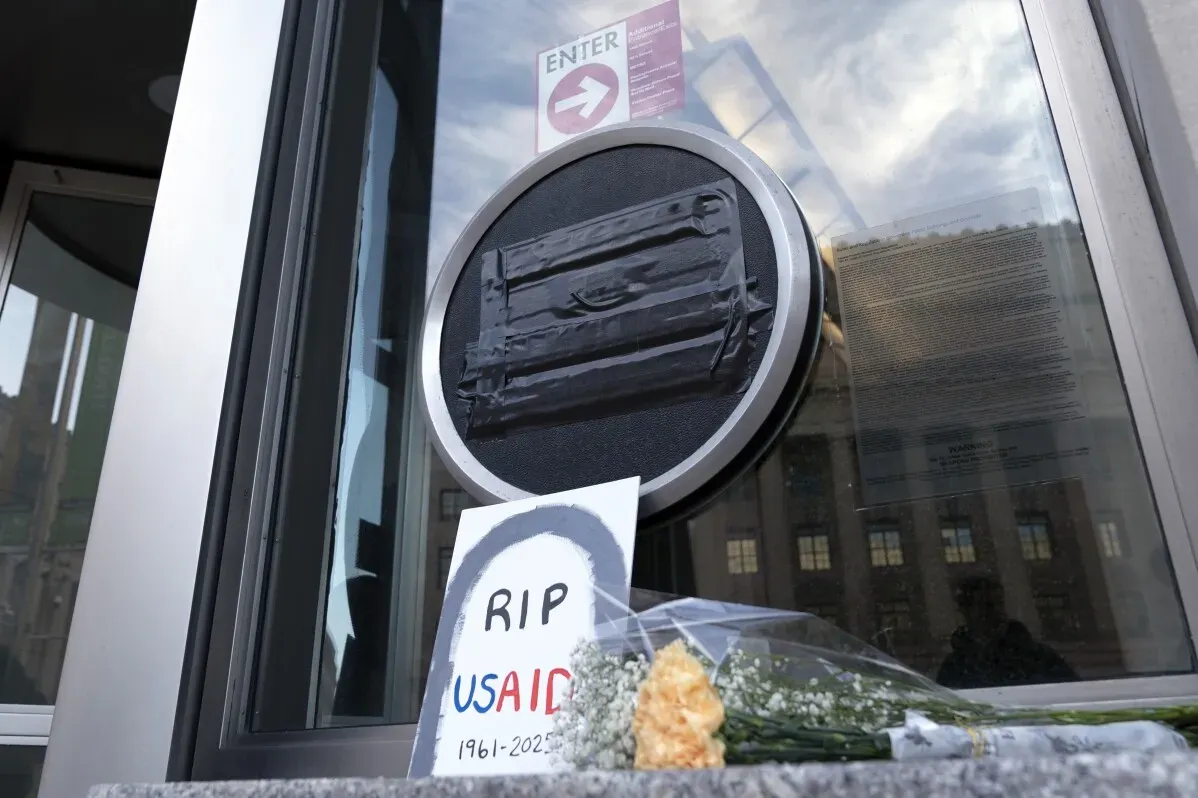 شعار وكالة USAID مغطى بشريط لاصق، مع لافتة "RIP USAID" وزهور عند مدخل المكتب، تعبيرًا عن الاحتجاج ضد تجميد المساعدات الخارجية.