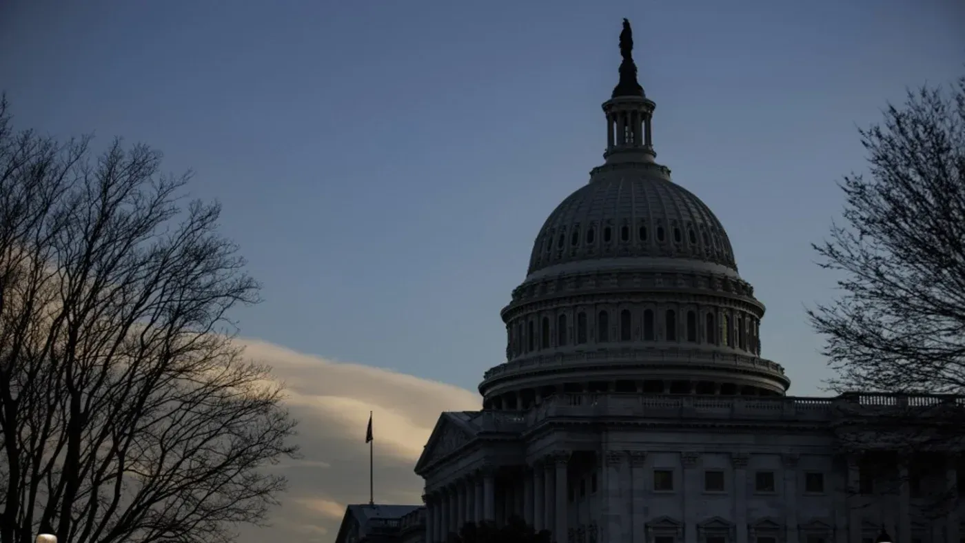 مبنى الكابيتول الأمريكي silhouetted against سماء غائمة، مع أشجار عارية في المقدمة، مما يعكس أجواء سياسية متوترة.