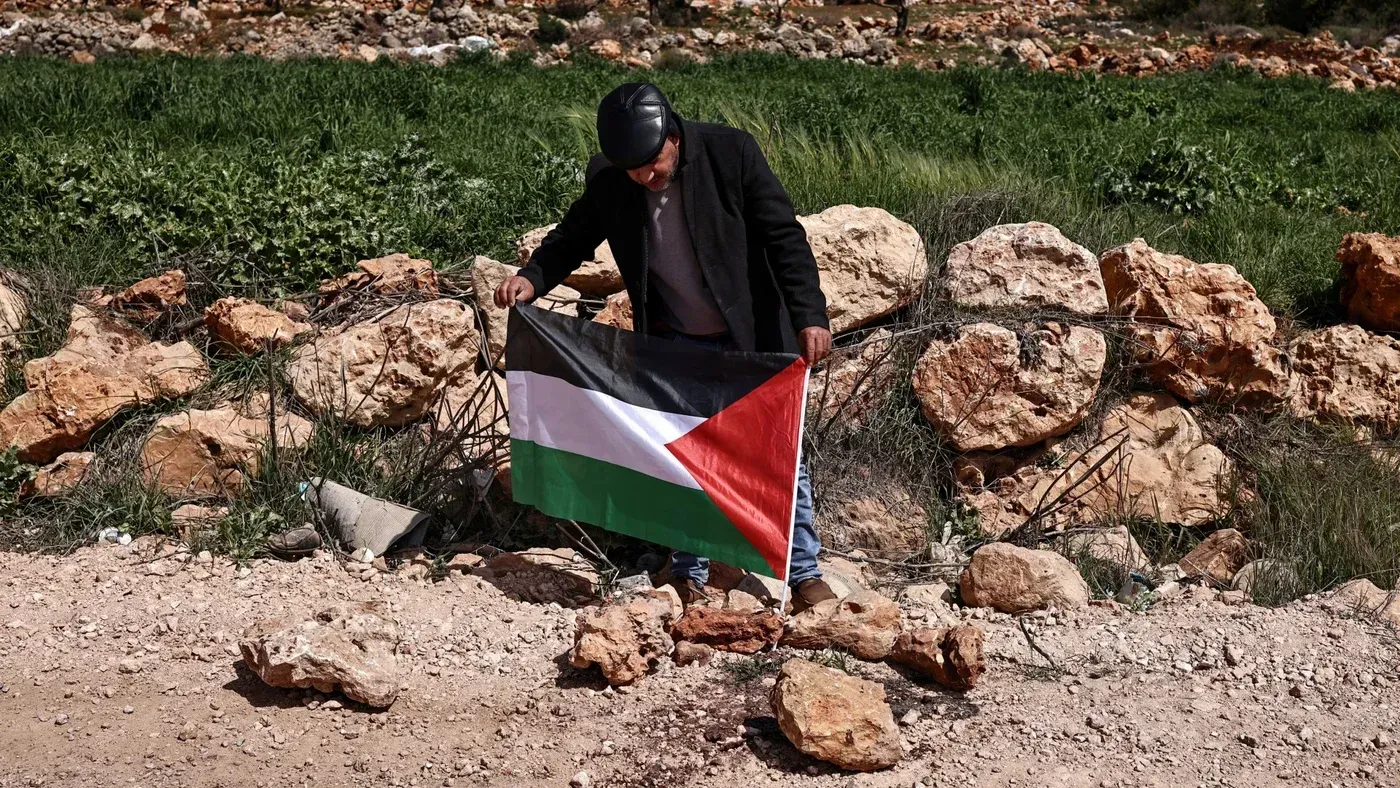 رجل يحمل علم فلسطين أمام صخور في منطقة خضراء، تعبيرًا عن التضامن مع الفلسطينيين في ظل تصاعد العنف من المستوطنين الإسرائيليين.