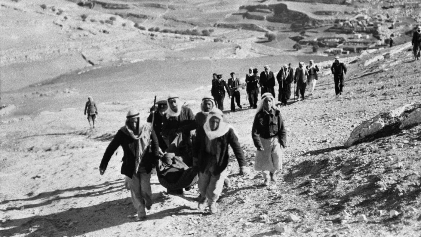 مجموعة من الرجال الفلسطينيين يحملون جثة على منحدر جبلي، بينما يسير آخرون في الخلف، تعكس آثار النكبة عام 1948.