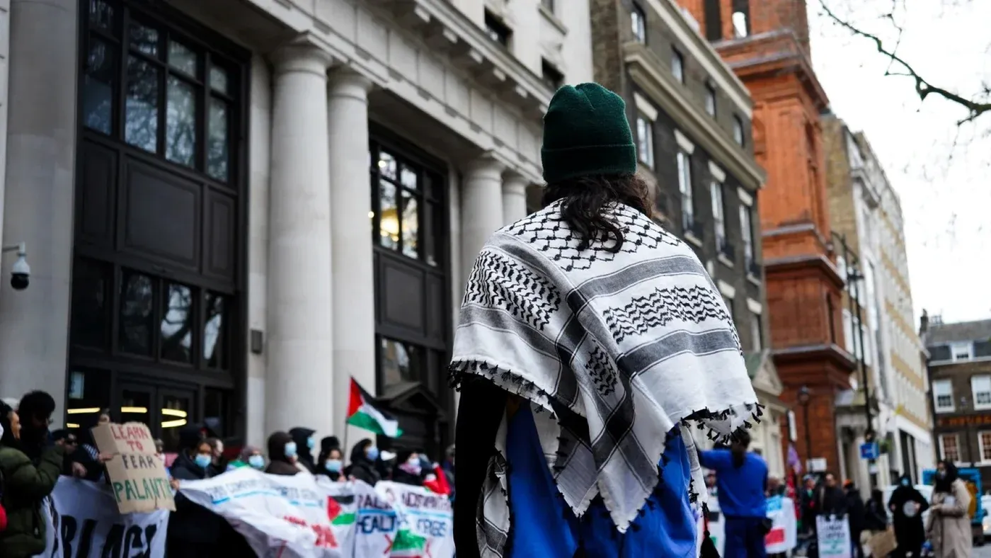 شخص يرتدي شالاً فلسطينياً يقف أمام حشد من المتظاهرين في لندن، يعبرون عن دعمهم لفلسطين خلال احتجاج.
