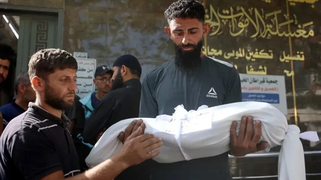 رجال يحملون جثمانًا ملفوفًا في قماش أبيض أمام مركز للتكفين في غزة، وسط أجواء من الحزن والتوتر.
