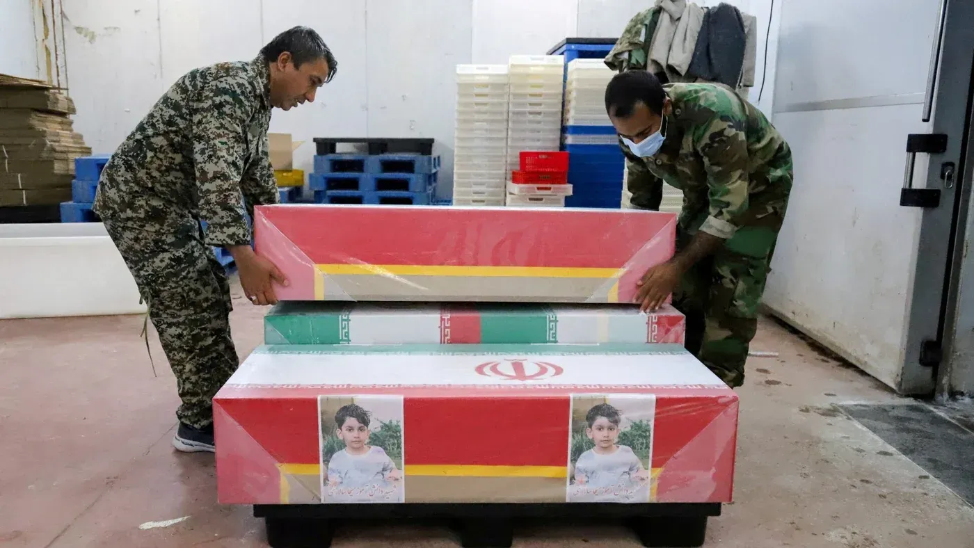 رجال يرتدون زيًا عسكريًا يقومون بحمل توابيت ملونة بأعلام إيران، تحمل صور أطفال، في مكان مخصص لتجهيز الجثامين بعد الهجوم على مدرسة البنات.