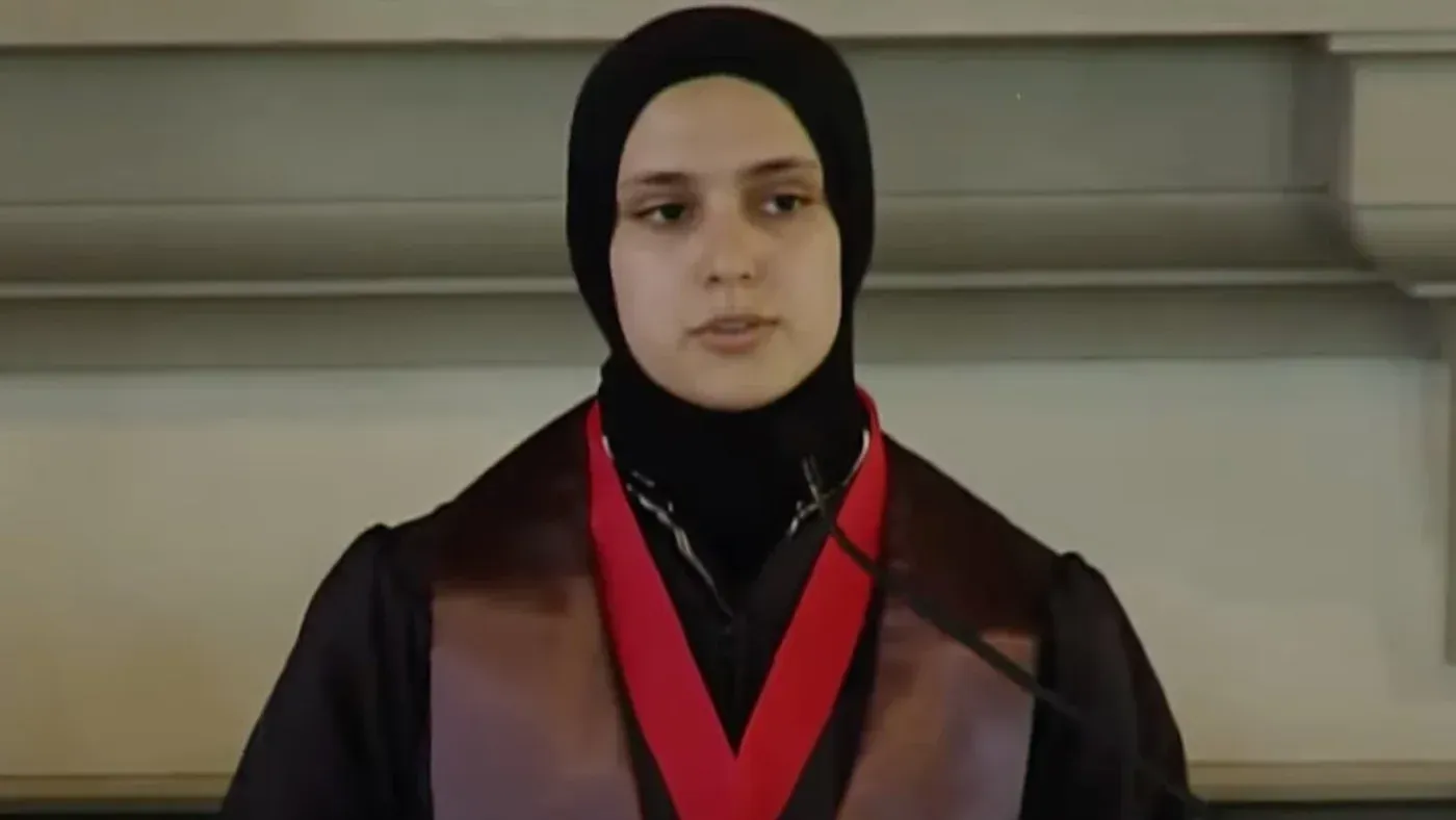 امرأة مسلمة ترتدي الحجاب وتلقي خطابًا في مناسبة رسمية، تعبر عن احتجاجها على قرار إقالة زميلتها من لجنة الحرية الدينية.