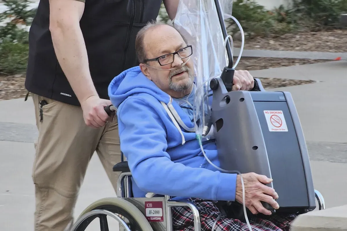 رجل مسن في كرسي متحرك يتلقى الأكسجين، يُنقل بواسطة شخص آخر، في سياق تأجيل جلسة استماع تتعلق بجرائم سرقة.