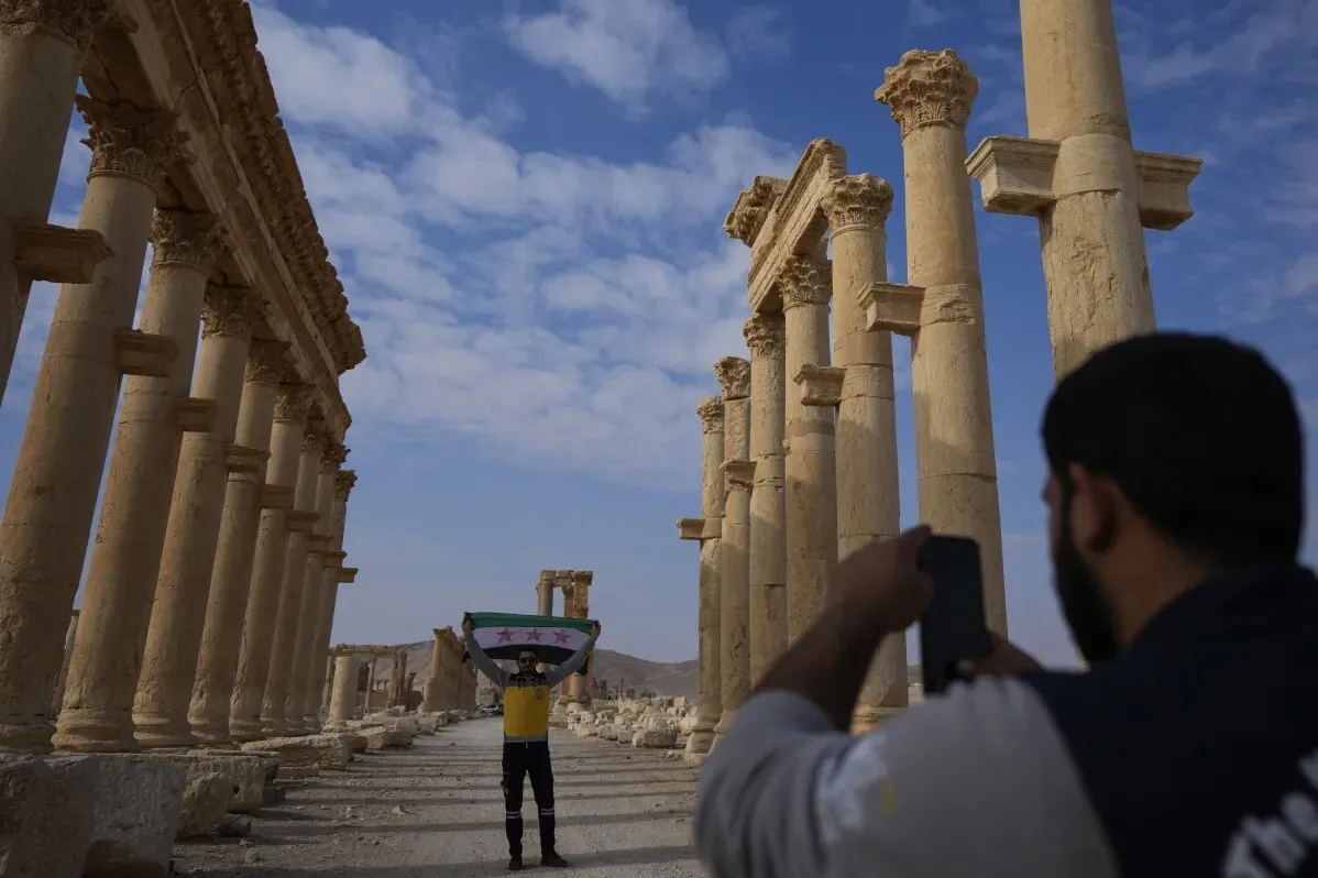 سائح يحمل علم سوريا يقف أمام الأعمدة الأثرية في تدمر، مع وجود زائر آخر يلتقط صورة له، مما يعكس جهود إحياء السياحة في الموقع التاريخي.