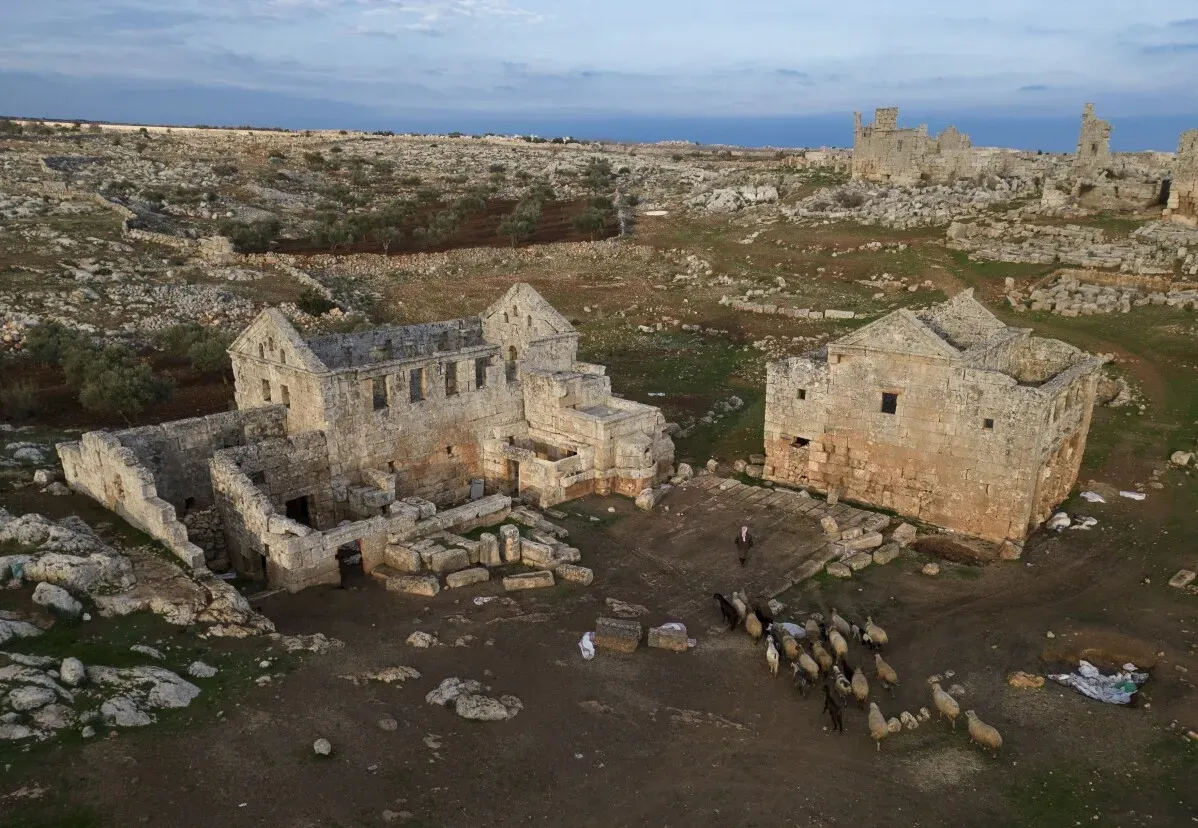 أنقاض معمارية تاريخية في كراك دي شوفالييه، سوريا، تظهر آثار الحرب، مع رعاة الغنم في الخلفية، تعكس أهمية التراث الثقافي المهدد.
