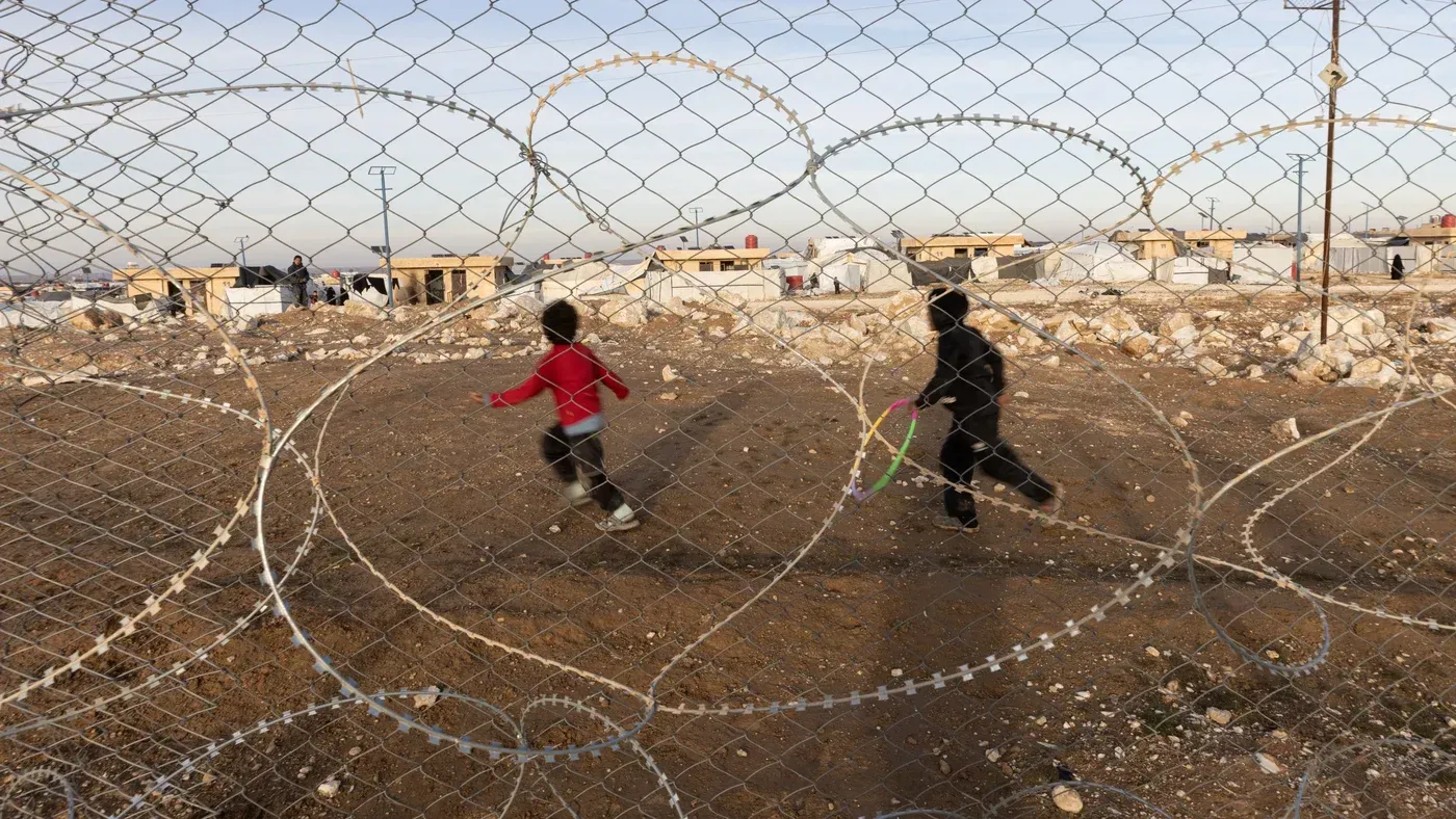 طفلان يلعبان في مخيم تحت سياج شائك، مما يعكس الوضع الصعب للأطفال المحتجزين في شمال شرق سوريا.