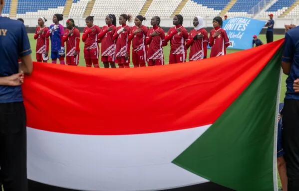 فتيات سودانيات يرتدين قمصانًا حمراء، يقفن في صف واحد على أرضية ملعب، يحملن علم السودان، يمثلن أول مشاركة دولية لمنتخب السيدات منذ الحرب الأهلية.