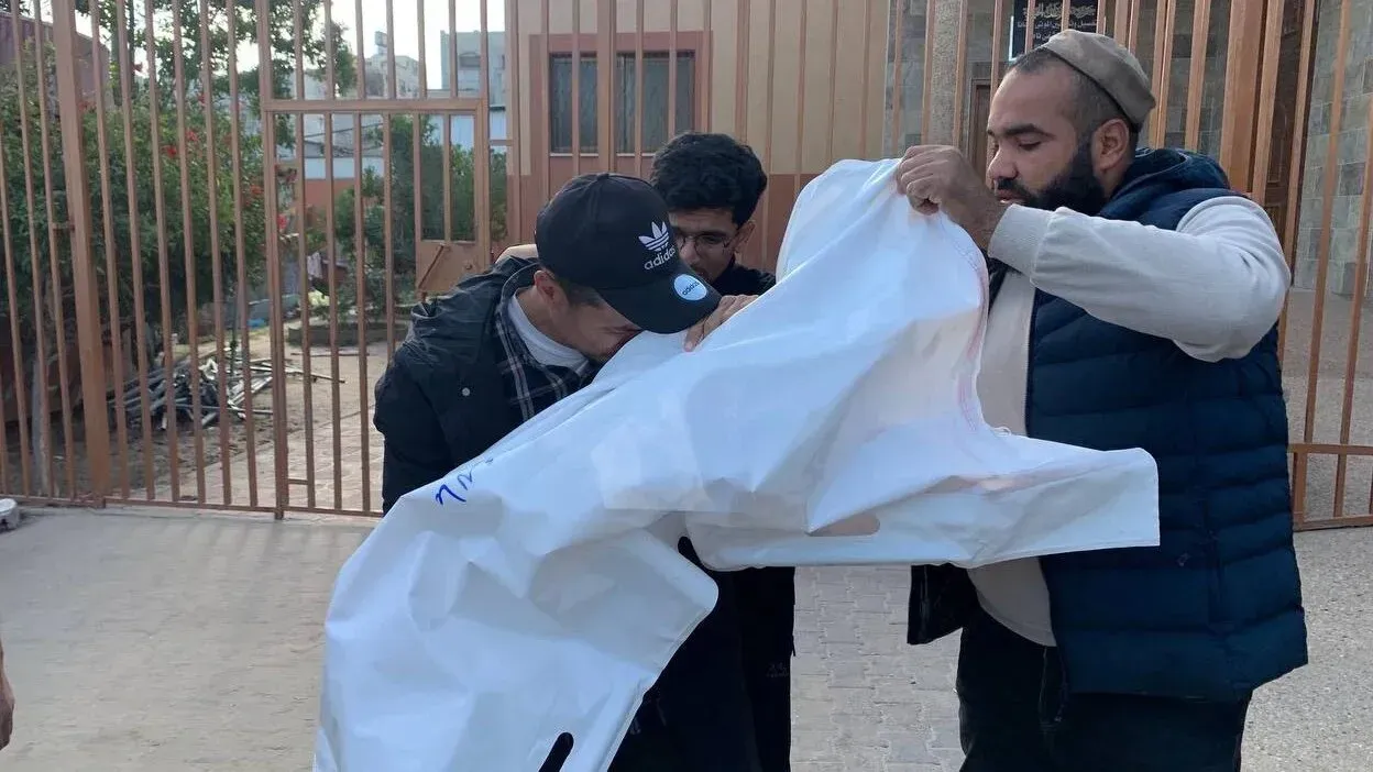 رجال يحملون كفنًا أبيض في منطقة مفتوحة، مع خلفية تظهر سياجًا حديديًا، تعبيرًا عن الحزن بعد فقدان عائلة في الهجوم على غزة.