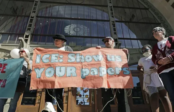 محتجون يحملون لافتة تطالب إدارة الهجرة والجمارك بإظهار الوثائق، أمام مبنى حكومي، مع تعبيرات تدل على القلق بشأن سياسات الهجرة.