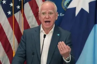 مسؤول حكومي يتحدث في مؤتمر صحفي حول حملة الهجرة في مينيابوليس، مع العلم الأمريكي في الخلفية، وسط توتر بعد حادثة إطلاق نار.