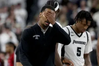 مدرب فريق بروفيدنس يبدو محبطًا بعد مشاجرة خلال مباراة ضد سانت جونز، بينما يظهر اللاعبون في الخلفية.