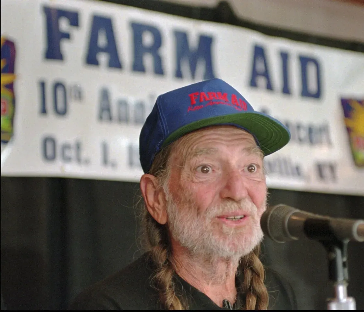 مؤسس "Farm Aid" ويلي نيلسون يتحدث في المؤتمر الصحفي العاشر للفعالية، مع لافتة تحمل اسم "Farm Aid" خلفه.