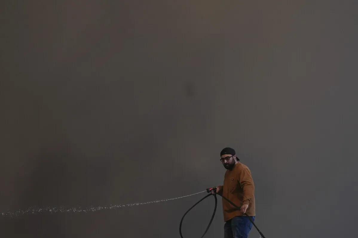 رجل يقوم بإخماد النيران باستخدام خرطوم مياه في منطقةaffected by Hughes Fire بكاليفورنيا، بينما تتصاعد الدخان في الخلفية.