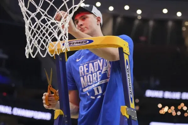 لاعب كرة سلة يقف على سلم ويقطع شبكة السلة بعد الفوز في بطولة NCAA، مرتديًا قميصًا أزرق وقبعة، مع أجواء احتفالية في الخلفية.