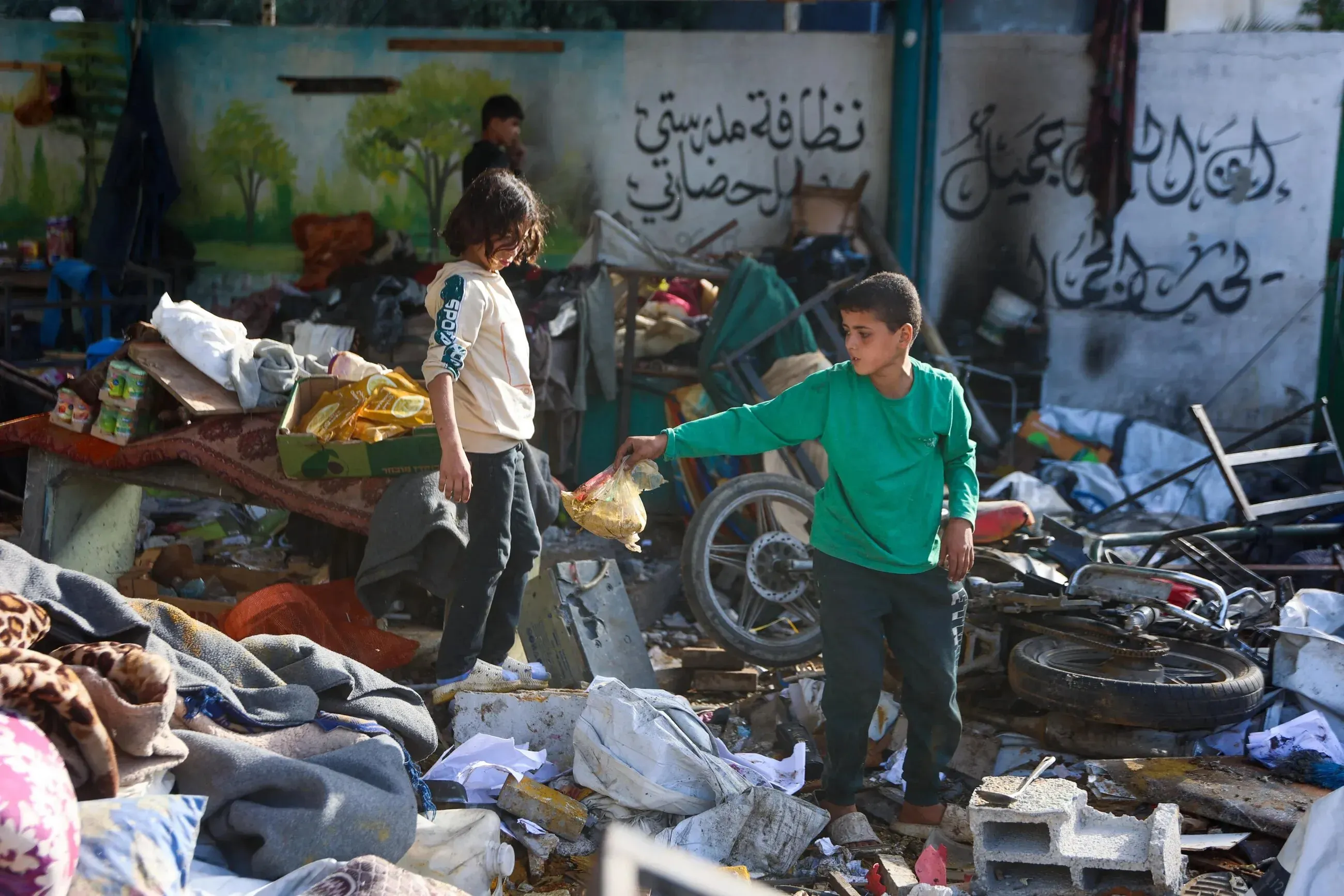 طفلان في مدرسة مدمرة بغزة، يتجولان بين الحطام والخراب بعد قصف إسرائيلي، مما يعكس الأثر المدمر للصراع.